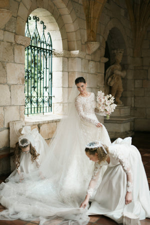 Cassandra Long Blusher Cathedral Veil shown in a dramatic, regal interior. Two flower girls arrange the sheer tulle train while the bride smiles, holding a white bouquet.