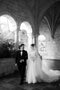 The Cassandra Long Blusher Veil covering the bride's face as she walks arm-in-arm with her father in a stone cloister.
