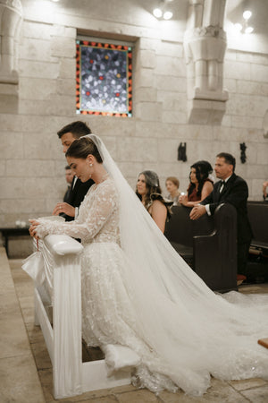 Bride wearing the Cassandra Veil with the long blusher pulled back and open, creating a voluminous two-tier silhouette. The sheer Italian tulle train cascades behind her as she kneels during the ceremony.