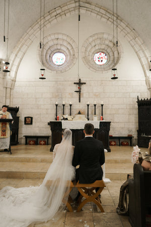 Colour photo of the Cassandra veil's pale ivory Italian tulle train pooling on the church floor, viewed from behind the bride and groom sitting at the altar during the ceremony.
