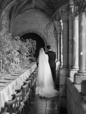 Bride and groom walking past the candlelit reception table in a stone cloister. The Cassandra veil elegantly pulls behind the bride.