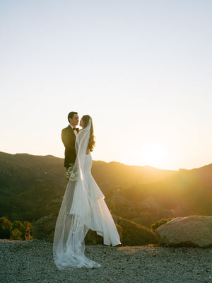 Bride and groom standing in the sunset with a mountain view; the bride is wearing a very long Dream Butterfly Veil trailing on the gravel.