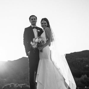 Black and white back lit portrait of a smiling bride wearing the long sheer Dream Butterfly Veil draped over her shoulder; her groom is beside her.