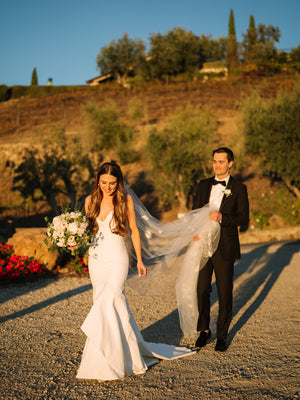 Bride wearing the Dream Butterfly Veil and fitted crepe wedding dress is walking on a gravel path while the groom holds the veil's train in a sunny vineyard setting.