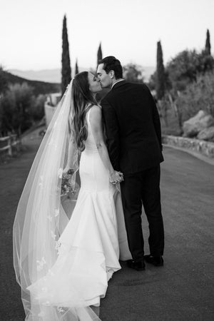 Black and white photo of a bride in a fitted dress and long sheer Dream Butterfly Veil kissing the groom on a winding road.
