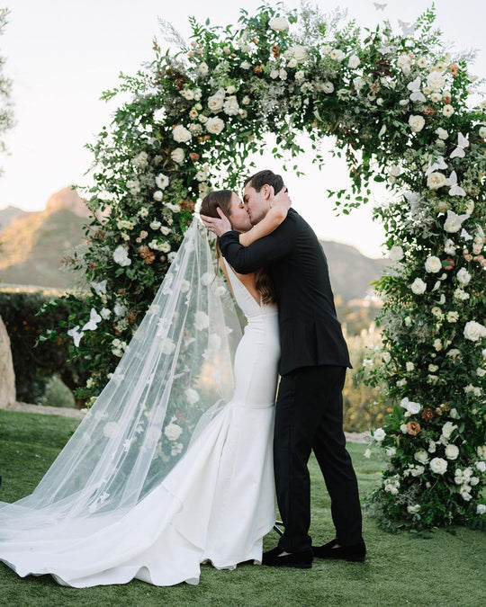 Bride wearing butterfly veil sharing first kiss with groom in front of floral arch.