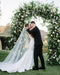 Full-length outdoor shot of the Dream butterfly veil, capturing the illusion tulle train flowing out behind the bride as she shares a kiss with her groom under a white floral arch.