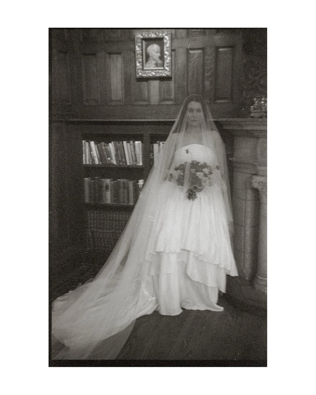 Black and white photograph of a bride in a formal setting with a bookshelf in the background.