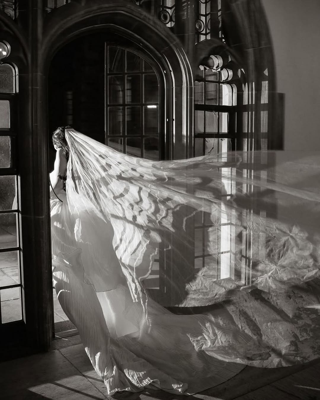 Bride wearing a very long silk wedding veil is existing an old church though an arched wood door.