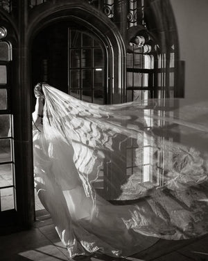 A dramatic black and white photograph of the Grace Pure Silk Veil on a bride as she exists through an arched doorway, the sweeping train of the veil is billowing out behind her. 