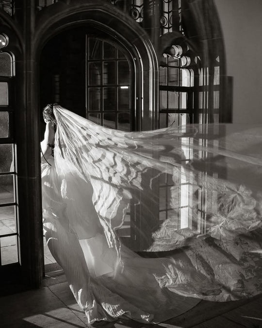 Bride wearing a very long silk wedding veil is existing an old church though an arched wood door.