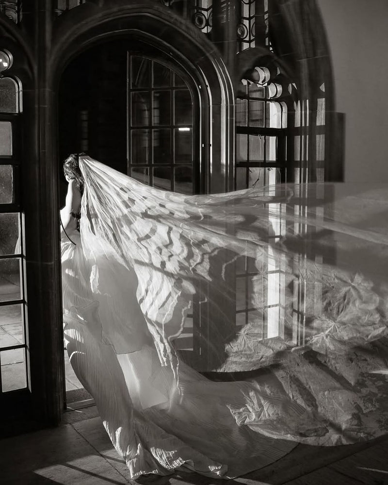 A dramatic black and white photograph of the Grace Pure Silk Veil on a bride as she exists through an arched doorway, the sweeping train of the veil is billowing out behind her. 