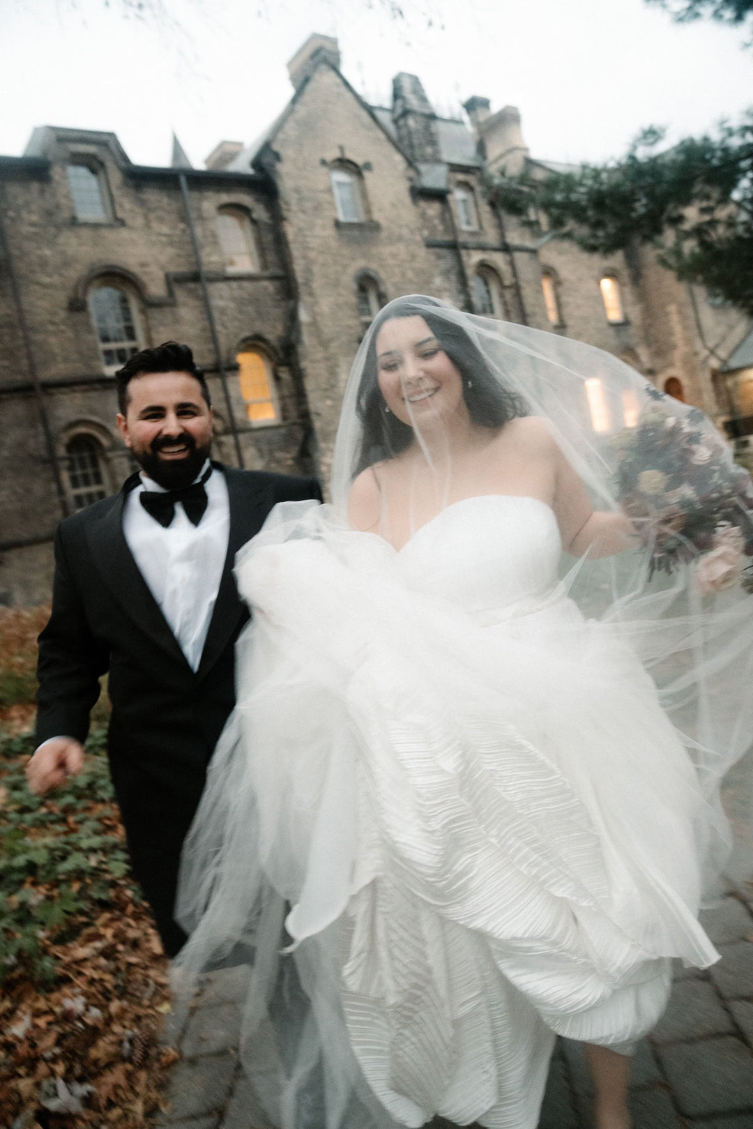 Couple in wedding attire standing in front of a stone building. The bride is wearing a Grace silk wedding veil from Noon on the Moon.