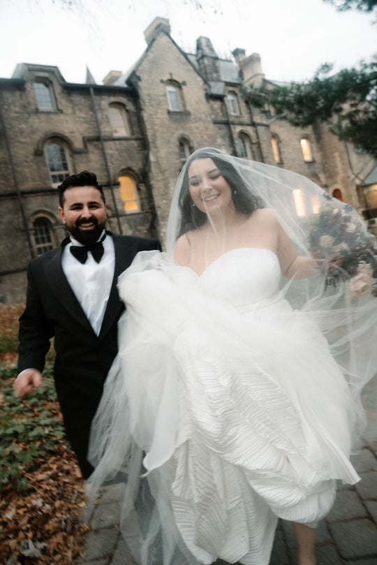 Couple in wedding attire standing in front of a stone building. The bride is wearing a Grace silk wedding veil from Noon on the Moon.