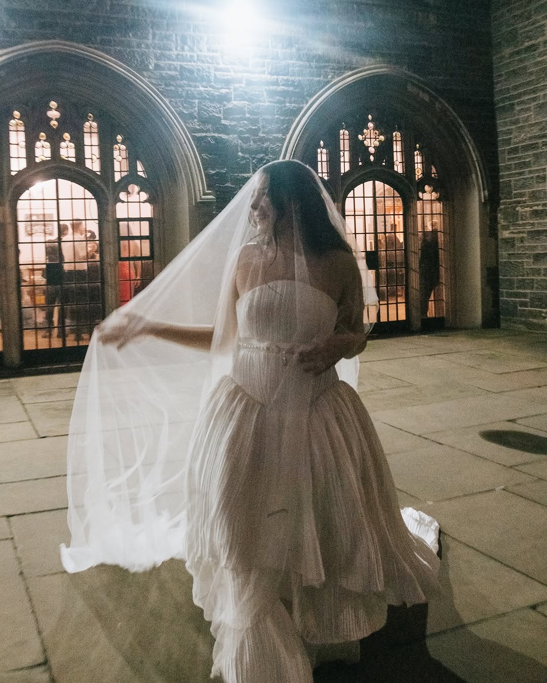 Bride wearing a long silk wedding veil and silk wedding dress is standing in front of a stone church wall with arched windows.