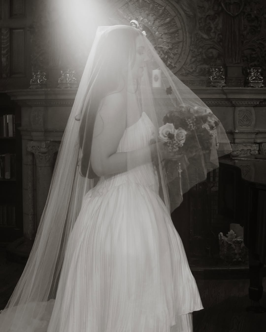 A bride wearing a long silk wedding veil is standing in an old church. She is holding the bouquet under the blusher.