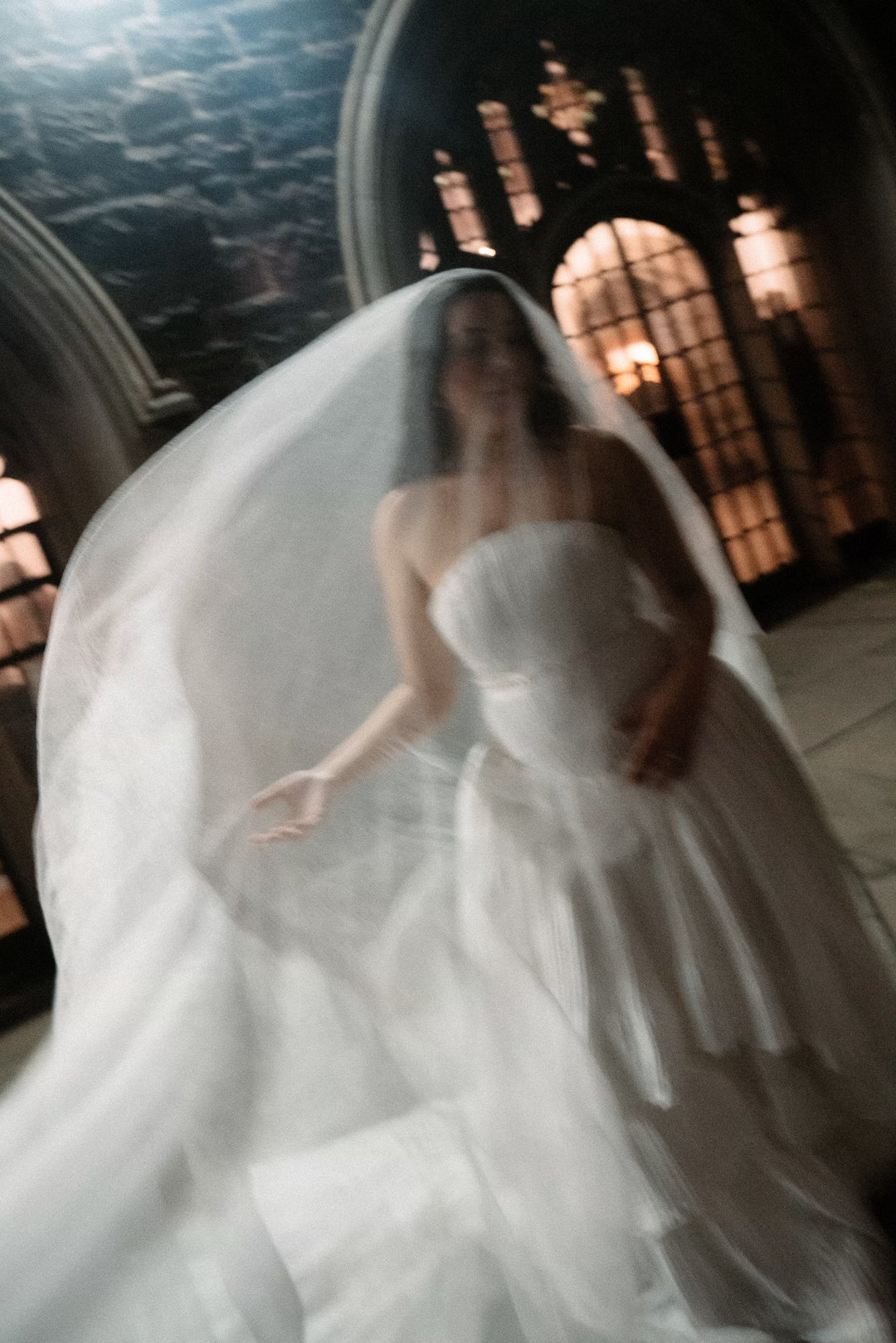 A bride wearing a long silk wedding veil and a silk wedding dress is standing in a dim stone church.