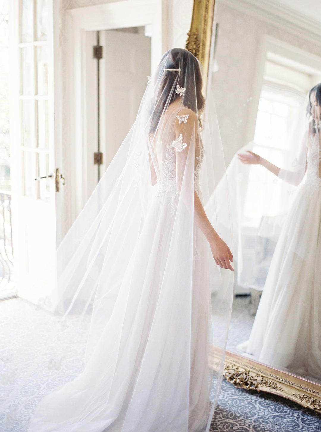 A bride wearing the REVERIE wedding veil with butterflies and lace wedding dress; she is standing in front of the mirror in the Graydon Hall bridal suite. 