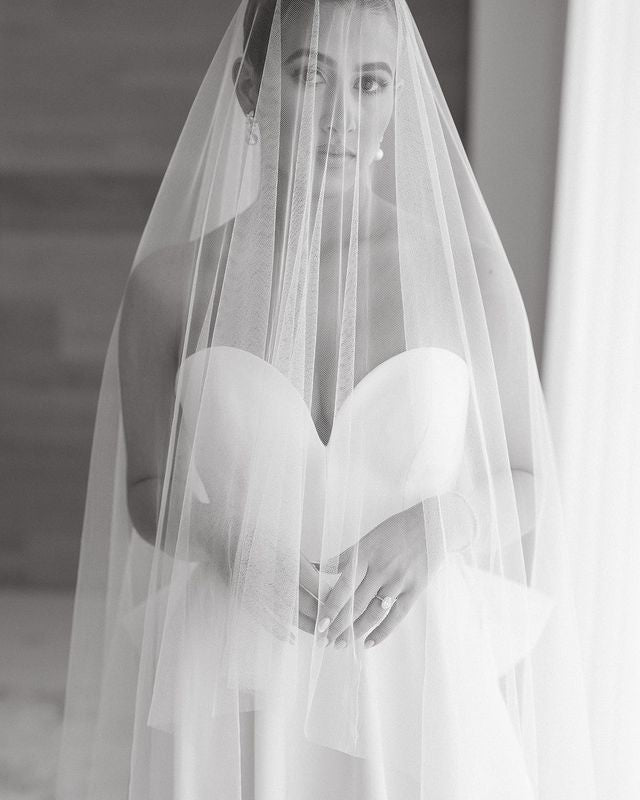 A black and white photograph of a bride wearing a soft wedding veil over her face. 
