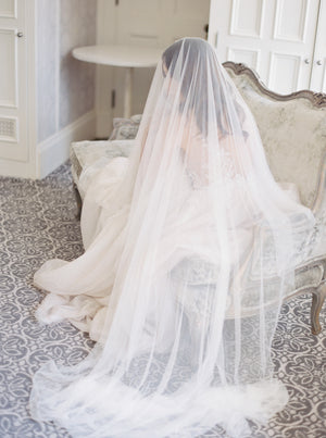 A full-length bridal portrait in Graydon Hall Manor. The bride is seated on an antique sofa, covered entirely by the soft and luxurious Angelique French Silk Drop Veil.