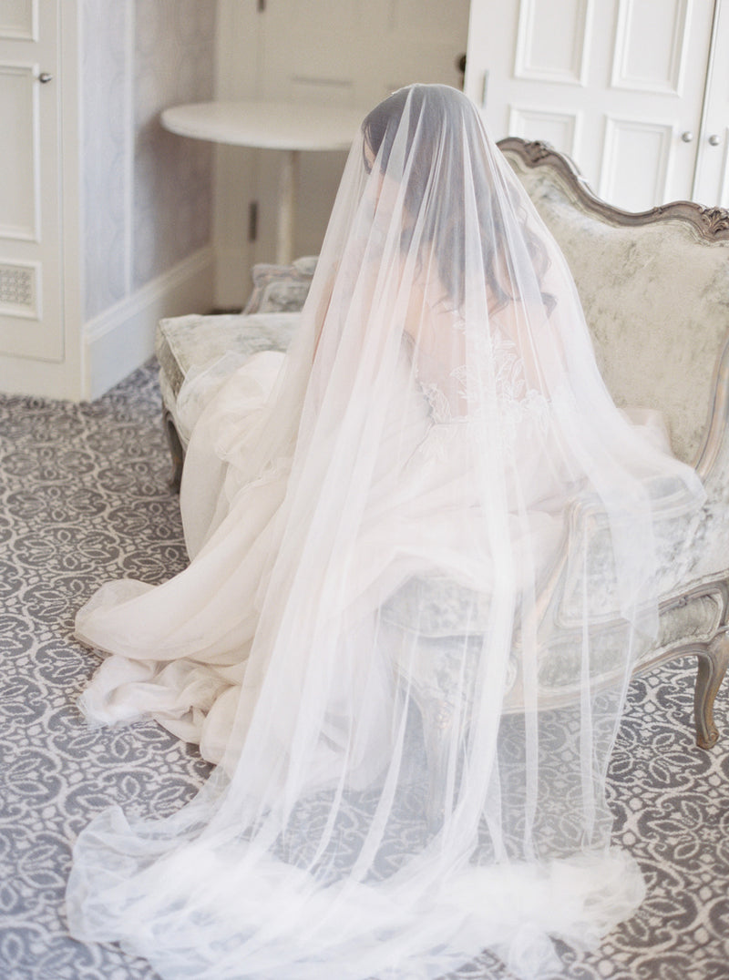 A full-length bridal portrait in Graydon Hall Manor. The bride is seated on an antique sofa, covered entirely by the soft and luxurious Angelique French Silk Drop Veil.