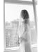 Black and white photo of a bride with a bob hairstyle standing by a window. The Angelique Silk Drop Veil drapes over her face, highlighting the veil's soft silk tulle material and showcasing the off-shoulder wedding gown.