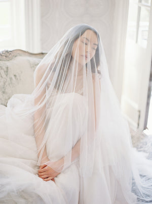 A romantic portrait of a bride sitting on an elegant velvet chair in the Graydon Hall manor bridal suite with the Angelique French silk drop veil worn over her face, draping softly, highlighting the fluid movement of the silk tulle.