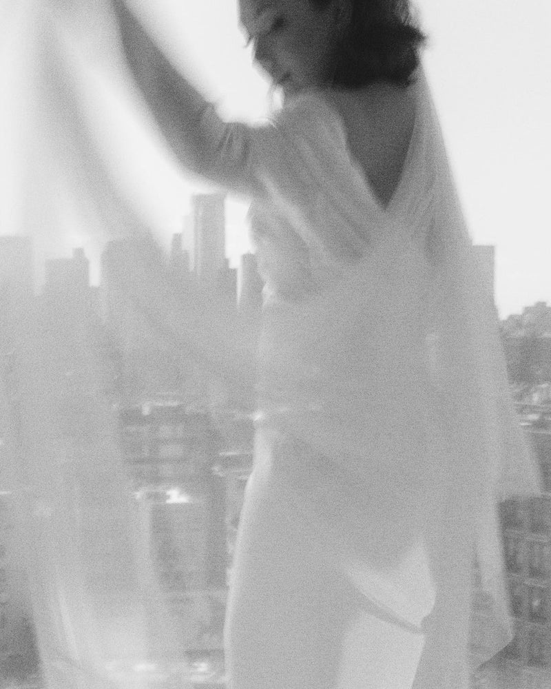 Blurred black and white photo of the bride dancing by a large window. The Angelique Silk Drop Veil flows and moves beautifully. 