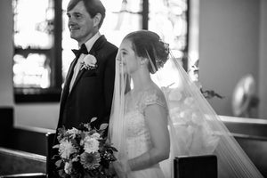 Black and white photo of a real bride walking down the aisle, smiling. The Angelique silk drop veil is worn over her face providing a classic, soft look.