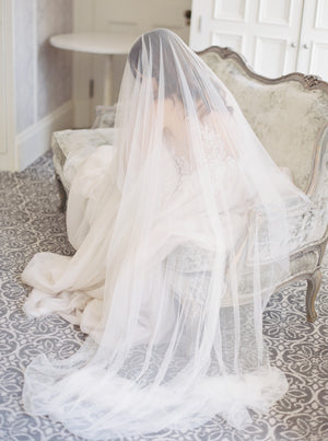 Full-length shot of a bride sitting on an antique sofa at Graydon Hall Manor. The Angelique French silk drop veil is draped over her and softly pools on the floor.