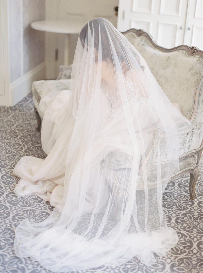 Full-length shot of a bride sitting on an antique sofa at Graydon Hall Manor. The Angelique French silk drop veil is draped over her and softly pools on the floor.