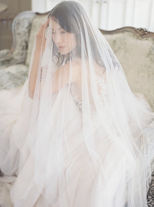 A veiled bride touches her hair while seated on an antique sofa in Graydon Hall Manor. The fluid Angelique French Silk Veil completely surrounds her in a soft layer. 