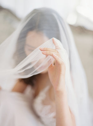 Extreme close-up of a bride lifting the sheer Angelique French Silk Drop Veil with her hand, showcasing the material's softness and light, elegant quality.