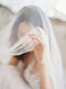 Extreme close-up of a bride lifting the sheer Angelique French Silk Drop Veil with her hand, showcasing the material's softness and light, elegant quality.