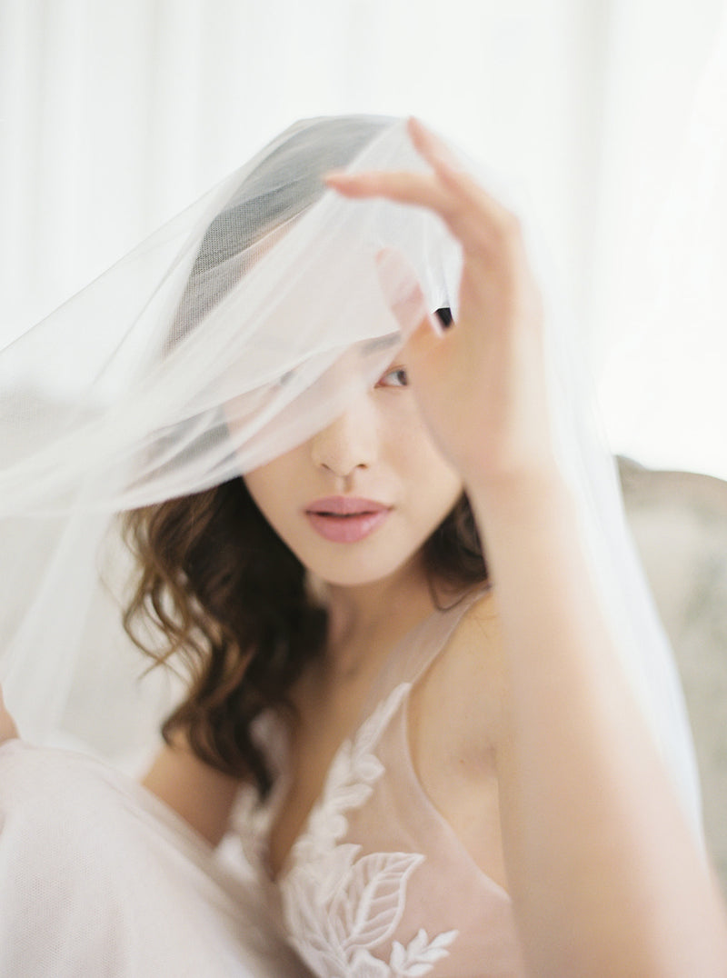 Close-up of a bride lifting the blusher of the Angelique drop veil to reveal her face. This detail shot emphasizes the luxurious fluidity of the silk tulle.