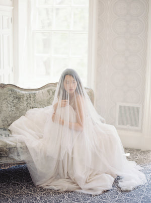 Romantic portrait of a bride in the light-filled Graydon Hall Manor bridal suite. She is veiled with the soft Angelique French Silk Drop Veil, seated on a velvet-covered chaise lounge.