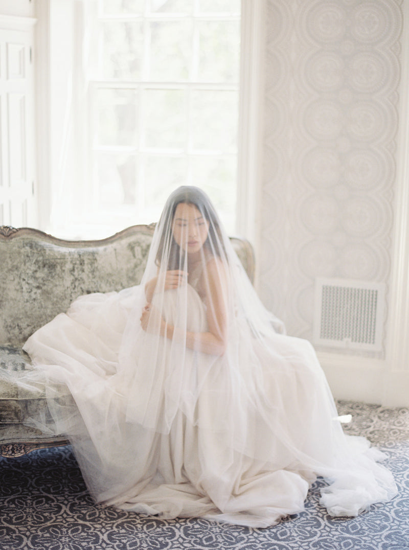 Romantic portrait of a bride in the light-filled Graydon Hall Manor bridal suite. She is veiled with the soft Angelique French Silk Drop Veil, seated on a velvet-covered chaise lounge.