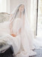 A romantic, softly lit photo of a bride sitting on a sofa in Graydon Hall Manor. The long, sheer Angelique French Silk Drop Veil softly covers her face.