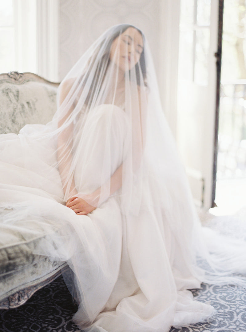 A romantic, softly lit photo of a bride sitting on a sofa in Graydon Hall Manor. The long, sheer Angelique French Silk Drop Veil softly covers her face.
