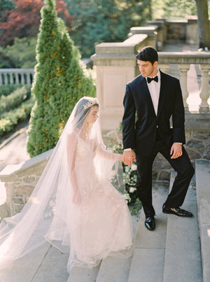 Bride and groom holding hands while walking down the grand stone stairs at Graydon Hall. The sheer Cassandra Veil in Imperial length drapes over the bride's gold leaf headpiece and lace gown.