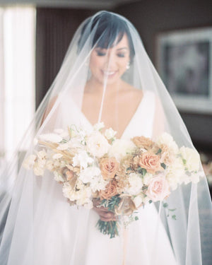 Real bride portrait wearing the Cassandra Veil's sheer long blusher draped over her face. The fine Italian tulle creates a beautiful, soft layer over the bride holding her warm-toned wedding bouquet.