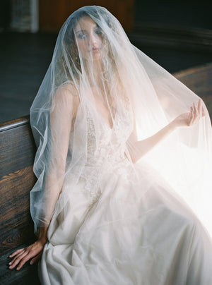 Close-up of bride sitting on a pew, pulling the veil over her face, showing the transparency over the gown neckline.