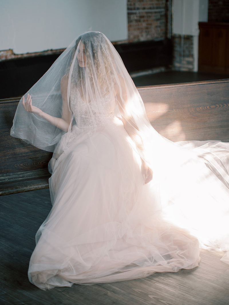 Bride sitting on a dark wooden pew with the Grace silk wedding veil pulled over her face, with sun streaming in, highlighting the sheer pure silk tulle.