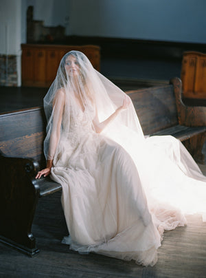 Bride wearing a soft tulle gown is sitting on a dark wooden pew, holding the Grace silk blusher veil to her face, creating a mysterious, romantic look.