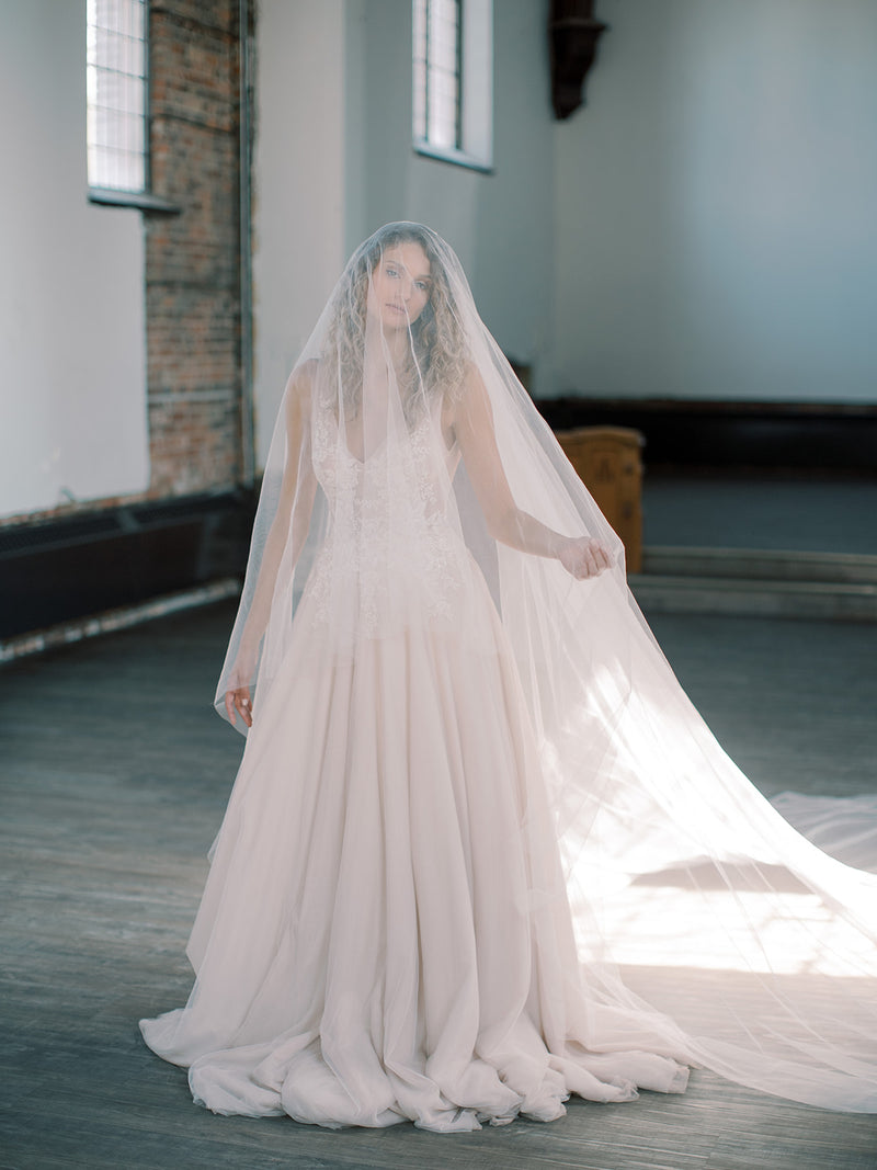 Full body shot of the bride standing in a high-ceiling church with high windows, with the Grace silk blusher veil covering her face and trailing dramatically behind her.