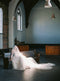 Full shot of the interior of a historic church. Bride sitting on a pew with the long train of the Grace silk blusher veil stretched out on the floor.