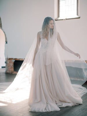 Bride wearing an a-line blush gown is standing in a sunlit church, side profile, showing the full length and width of the sweeping veil train.
