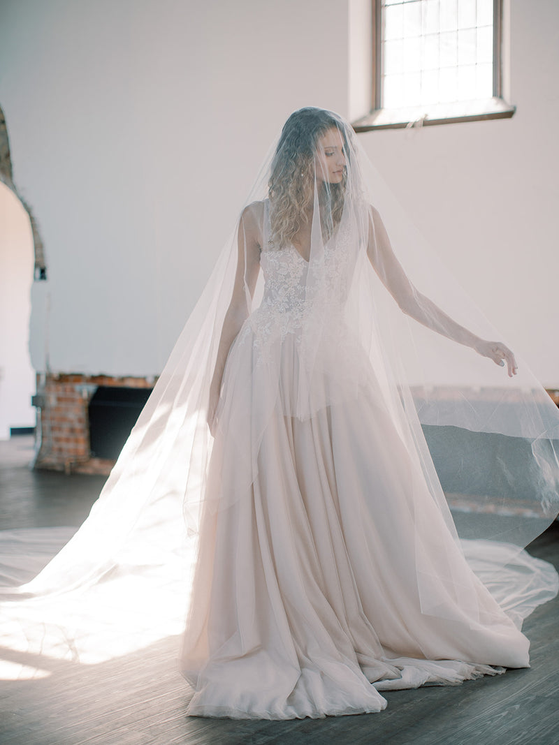 Bride wearing an a-line blush gown is standing in a sunlit church, side profile, showing the full length and width of the sweeping veil train.