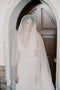 Straight-on portrait of the bride standing under an archway wearing the Grace Silk Blusher Veil with the sheer silk tulle draped over her face.