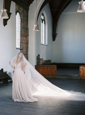 Wide shot of the bride standing alone in a large, bright church, showing the full, dramatic length of the Grace silk wedding veil train laid out.